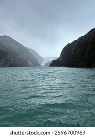 Dramatic view of a glacier surrounded by steep mountains and a serene lake under a cloudy sky