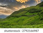 A dramatic view of Glen Ogle Viaduct crossing the steep hillside of Glen Ogle near Lochearnhead in the Scottish Highlands. The historic stone railway viaduct curves gracefully through vibrant green slopes beneath a glowing evening sky, surrounded by rugged mountains, woodland and rolling Highland scenery.