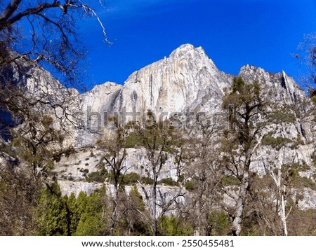 Dramatic view of a granite mountain with a waterfall near the Merced Riiver in Yosemite National Park with lush evergreen forest in the foreground and blue sky in the background.