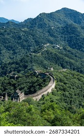 A dramatic view of the Great Wall of China winding across steep, lush green mountains under a clear sky. The scene highlights the massive scale of the historic structure and natural landscape.