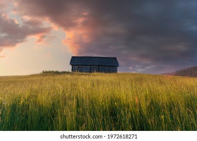 Dramatic View Of High Grass Mountain Hill Under Sunset Clouds With The Old Wooden Rustic House. Summer Landscape.