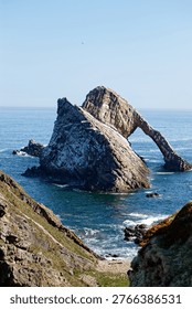 Dramatic view of the iconic Bow Fiddle Rock, a natural sea arch located on the Moray coast near Portknockie, Scotland, under a clear blue sky.