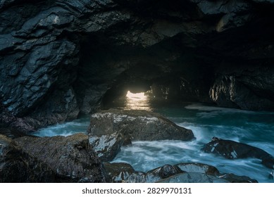 A dramatic view from inside a rugged ocean cave looking outward toward the open sea, with natural rock formations framing the horizon and waves moving across the water beyond. The dark, textured cave interior contrasts with the bright blue ocean outside, creating a strong sense of depth and perspective. Natural light enters through the cave opening, highlighting the weathered rock surfaces and capturing the secluded, raw beauty of the coastline.