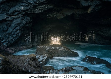 A dramatic view from inside a rugged ocean cave looking outward toward the open sea, with natural rock formations framing the horizon and waves moving across the water beyond. The dark, textured cave interior contrasts with the bright blue ocean outside, creating a strong sense of depth and perspective. Natural light enters through the cave opening, highlighting the weathered rock surfaces and capturing the secluded, raw beauty of the coastline.