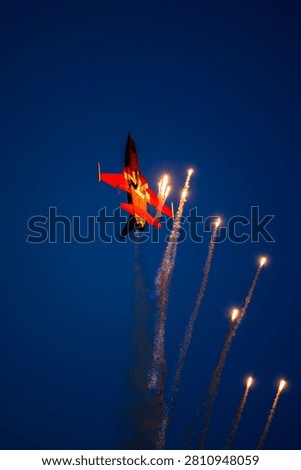 A dramatic view of an inverted fighter jet performing a nighttime maneuver, firing dazzling pyrotechnic flares against a deep twilight blue sky during