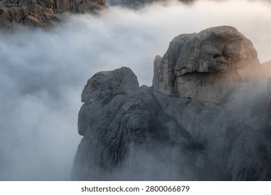 A dramatic view of jagged mountain peaks rising above a dense layer of low-hanging clouds during a sunrise.