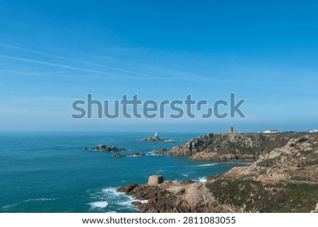 Dramatic view of La Corbiere Lighthouse on rugged cliffs with turquoise sea, rocky islets and clear blue sky, evoking calm and maritime history. La Corbiere, Saint Brelade, Jersey, UK.