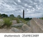 A dramatic view of the Laboe Naval Memorial in Schleswig-Holstein, Germany, captured under cloudy skies with a foreground of sandy dunes, grasses, and rocks.