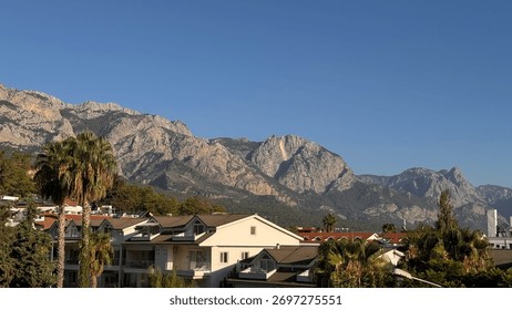 Dramatic view of large, rocky mountains towering over white Mediterranean residential buildings and lush green palm trees under a clear blue sky
