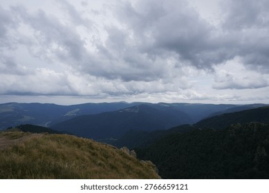 A dramatic view of layered mountain ranges under a heavy, overcast sky, with a foreground of dry, golden grass on a windswept hill.