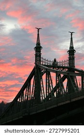  A dramatic view of the Liberty Bridge (Szabadság híd) in Budapest captured at sunset, with its distinctive green iron structure and decorative towers silhouetted against a vivid pink and blue evening sky. The textured clouds and warm twilight colors create a cinematic, atmospheric mood that highlights the historic architecture spanning the Danube River. This vertical image offers strong visual impact and generous copy space, making it ideal for designers, graphic artists, and editorial or web projects focused on travel, urban identity, architecture, and atmospheric city backgrounds.