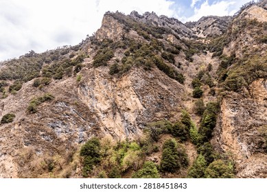 Dramatic view looking directly up at a massive, sheer mountain cliff covered in rocky outcroppings and patchy green forests under a cloudy sky in Yunnan.