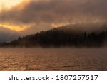 Dramatic view of a natural landscape in a mountainous area with a quiet lake and a coniferous forest shrouded in fog with the sun rising behind the clouds in the early hours of the morning.