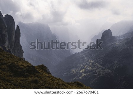 Dramatic view of Picos de Europa mountains with jagged cliffs, deep valleys, and misty atmosphere as sunlight breaks through thick clouds over the rugged autumn landscape