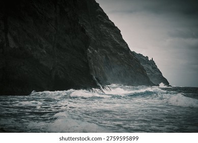Dramatic view of powerful ocean waves crashing against a majestic, dark rocky cliff under a stormy, overcast sky. A wild and untamed natural landscape showcasing the raw power of the sea.