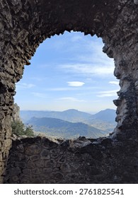Dramatic view of the Pyrenean mountain ranges seen through a centered oval stone opening in the dark basalt walls ofChâteau de Peyrepertuse, one of the most spectacular Cathar castles in southern France. The ancient stone arch acts as anatural frame, creating a striking composition with the blue sky and high-altitude wispy clouds beyond. A powerful blend ofmedieval military architecture and wild mountain scenery in the Corbières region, Occitanie.