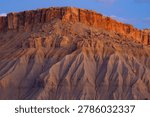 A dramatic view of a reddish-orange sandstone cliff contrasting with eroded gray soil structures under a clear blue sky.