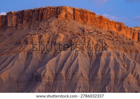A dramatic view of a reddish-orange sandstone cliff contrasting with eroded gray soil structures under a clear blue sky.