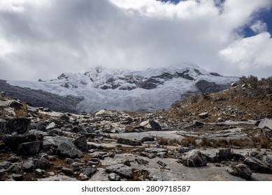 A dramatic view of a rocky, barren foreground leading toward a massive snow-capped mountain peak under a cloudy sky.