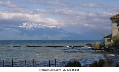 dramatic view of rough lake waves crashing against a concrete pier with a majestic snow-capped mountain rising under a cloudy sky