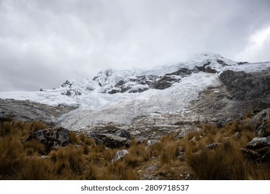 A dramatic view of a rugged glacial mountain peak surrounded by rocky terrain and dry alpine grasses under a cloudy sky.