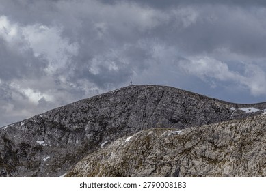 Dramatic view of a rugged mountain peak under a cloudy sky. A simple wooden cross stands prominently on the summit, silhouetted against the grey backdrop. The rocky terrain is partially covered with snow and sparse vegetation. The scene evokes a sense of solitude and resilience in a harsh natural environment. Perfect for representing themes of faith, perseverance, and the beauty of the alpine landscape.