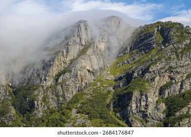 Dramatic view of rugged mountain peaks partially obscured by low-hanging clouds and mist, with steep rocky slopes and patches of vibrant green vegetation.