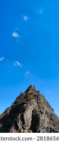 A dramatic view of a rugged mountain summit reaching towards a vast expanse of bright blue sky dotted with wispy white clouds.