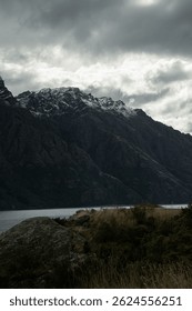 A dramatic view of rugged mountains dusted with snow under a moody, overcast sky. The scene overlooks a tranquil lake bordered by rocky terrain and wild grasses, evoking a remote, natural solitude.