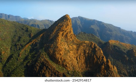 A dramatic view of a rugged, sunlit mountain peak with surrounding green-covered cliffs under a clear blue sky. - Powered by Shutterstock