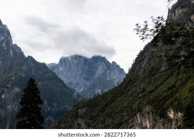 A dramatic view of rugged, towering mountain peaks in Yunnan, China, partially shrouded in thick, low-hanging clouds under an overcast sky.