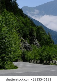 A dramatic view of a sharp bend on the Ossetian Military Road (Transcaucasian Highway) cutting through the Alagir Gorge in North Ossetia. The asphalt road curves in a tight arc around a sheer, rocky slope covered in dense green forest and shrubs. Along the right side of the road, concrete safety barriers with characteristic black-and-white warning markings guide drivers through this hazardous mountain passage. In the background, the majestic silhouettes of the Caucasus Mountains rise, their foothills wrapped in low white clouds that drift through the valley. The contrast between the man-made road infrastructure and the raw, untamed mountain landscape creates a powerful image of human engineering meeting nature's grandeur.
