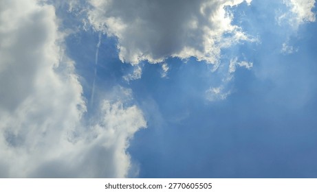 A dramatic view of the sky featuring thick, fluffy clouds partially covering the sun, with bright sun rays piercing through and creating a striking contrast against the vivid blue background. The interplay of light and shadow adds depth and a serene yet powerful atmosphere, symbolizing hope, clarity, and natural beauty. Ideal for themes related to weather, spirituality, inspiration, and nature backgrounds.