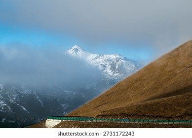 A dramatic view of a snow-capped Himalayan peak partially shrouded in mist, with a green-guardrailed mountain road curving along arid golden slopes on the Tibetan Plateau, China.