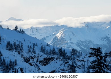 A dramatic view of snow-capped peaks engulfed in clouds, surrounded by rocky ragged ridges and slopes from Whistler Mountain, BC, Canada.