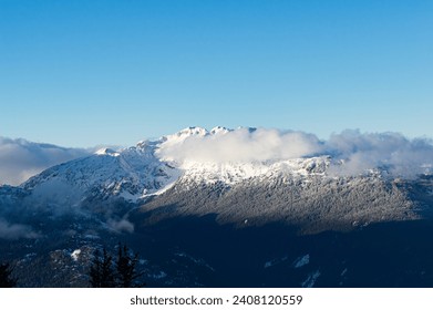 A dramatic view of snow-capped peaks engulfed in clouds, surrounded by thick forest, against a blue sky from Whistler Mountain, BC, Canada.