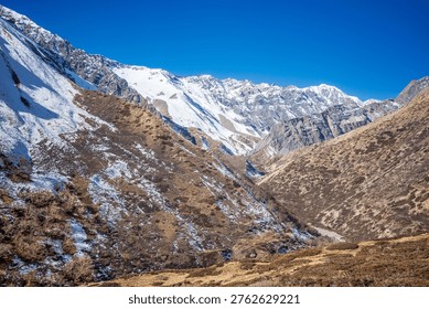 Dramatic view of a snow-covered Himalayan mountain range rising above a rugged alpine valley with dry high-altitude terrain and scattered snow patches. The steep slopes and layered ridgelines