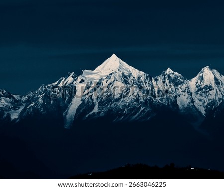 Dramatic view of a snow-covered Himalayan mountain peak under a dark blue sky. The majestic, icy summit and rugged ridges create a powerful and serene natural landscape ideal for winter themes.
