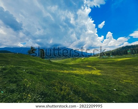 A dramatic view of snow-streaked mountain slopes beneath heavy storm clouds, with a wire fence, wind-swept trees, and small figures standing on a grassy overlook.