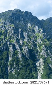 A dramatic view of steep, rocky mountain terrain with dense evergreen vegetation clinging to its surfaces under a partly cloudy sky