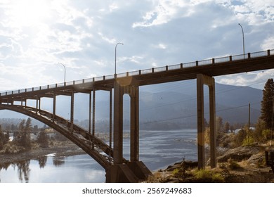 Dramatic view of a tall bridge spanning over a calm river with a backdrop of trees, mountains, and a bright sky.