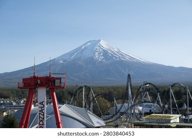 A dramatic view of a theme park with towering roller coaster tracks and a red ride structure in the foreground, set against the snow‑capped Mount Fuji under a clear blue sky. The contrast between the dynamic amusement attractions and the iconic natural landmark creates a striking and energetic scene.
