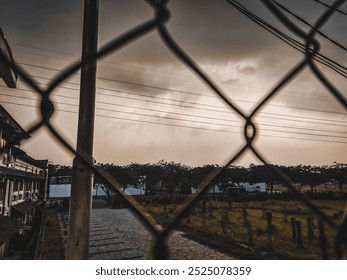 A dramatic view through a diamond-shaped wire fence, highlighting a vibrant sky with clouds illuminated by the setting or rising sun, casting a warm glow.