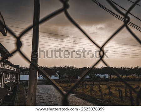 A dramatic view through a diamond-shaped wire fence, highlighting a vibrant sky with clouds illuminated by the setting or rising sun, casting a warm glow.