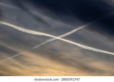 dramatic view of two distinct, bright white jet vapor trails contrails that intersect to form X shape. The background is a gradient sky from dark blue to warm orange sunset tones