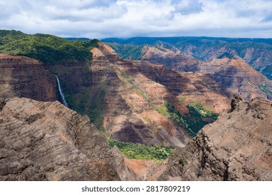 Dramatic view of Waimea Canyon featuring a tall thin waterfall cascading down colorful red and orange layered cliffs with green vegetation under a partly cloudy sky.