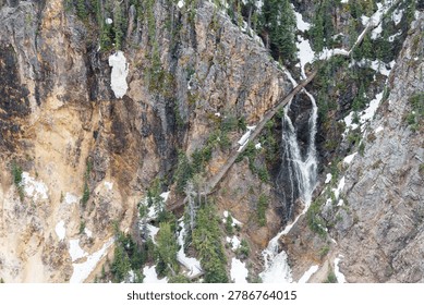 Dramatic view of a waterfall cascading down a rugged canyon with snow, green trees, serene mood