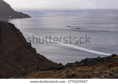 Dramatic volcanic cliffs overlook calm ocean waters with aquaculture fish farm nets visible offshore. Moody overcast coastal landscape in