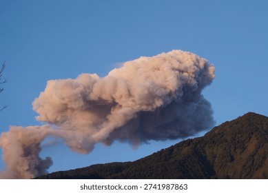 Dramatic volcanic eruption producing a massive ash cloud above a mountain landscape under clear blue sky. Natural disaster concept, environmental hazard, and geological phenomenon.                        