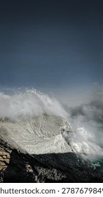 Dramatic volcanic landscape featuring textured sulfur cliffs partially covered by heavy white smoke and toxic gases at Ijen crater.